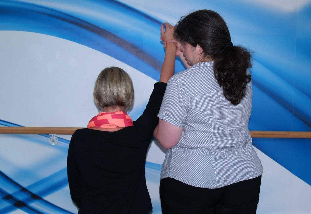 Zwei Frauen stehen vor einer Wand mit blauen Mustern; die eine hält den erhobenen Arm der anderen, als wolle sie bei einer Dehnungs- oder Rehabilitationsübung helfen.